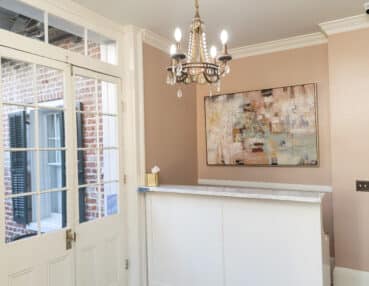 A small reception area with a white desk, abstract painting on the wall, chandelier, and glass-paned door leading outside.