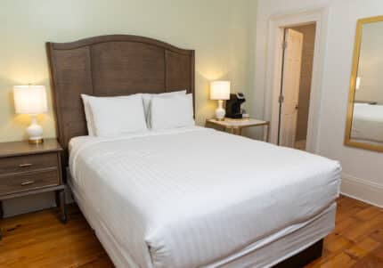 A neatly made bed with white linens and pillows in a room, flanked by wooden nightstands with lamps, a large mirror on the right wall, and an open door leading to another room.