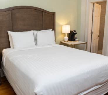 A neatly made bed with white linens in a room with soft lighting. A wooden headboard and nightstand are on the left, and a mirror reflects part of the room on the right.