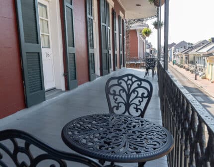 A wrought iron table and chairs set on an ornate balcony overlooking a street lined with colorful buildings under a clear sky.