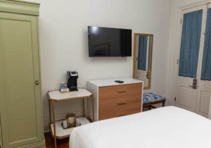 Hotel room with a bed, dresser, wall-mounted TV, mirror, green wardrobe, coffee maker on a small table, and blue cushioned bench by the door.