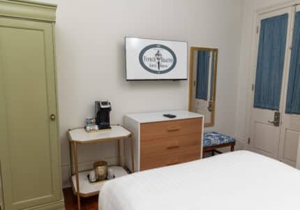 Hotel room with a bed, a green wardrobe, a dresser with a TV above it, a coffee maker on a small table, a wall mirror, and a bench by curtained doors.