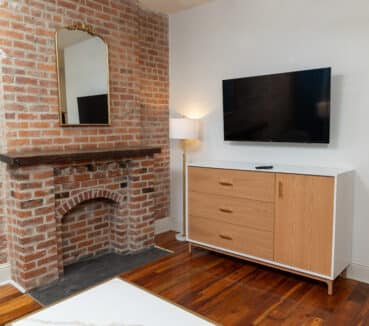 Room with exposed brick fireplace, wall-mounted TV, wooden dresser, and lamp.