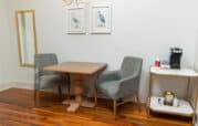 Two gray chairs and a wooden table sit near a wall with bird art. A gold-framed mirror is on the left. A small cart with a coffee maker and supplies is on the right.