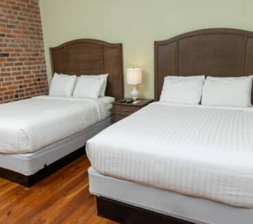 Two neatly made double beds with wooden headboards in a room with a brick wall and wooden floor. A small nightstand with a lamp is between the beds.