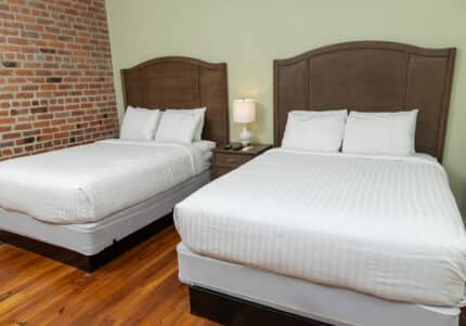 Two neatly made double beds with wooden headboards in a room with a brick wall and wooden floor. A small nightstand with a lamp is between the beds.