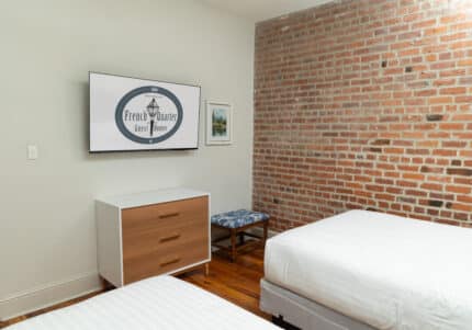 A bedroom with a brick wall featuring a wall-mounted TV displaying "French Quarter". There's a dresser, a picture frame, an upholstered bench, and two white beds.