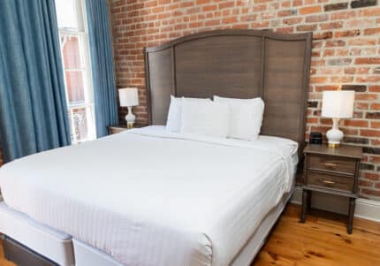 A neatly made bed with white linens and four pillows is flanked by two bedside tables, each with a lamp. The room features exposed brick walls and blue curtains by a window.