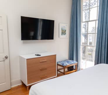 Bedroom interior with a wall-mounted TV above a wooden dresser, a framed picture, and a bench with a patterned cushion near tall windows with blue curtains and a red brick accent wall.
