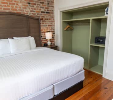Hotel room with a neatly made bed, wooden headboard, and a nightstand with a lamp. Open closet with hangers, an iron, and a safe. Exposed brick wall and wooden floor.