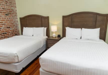 Two neatly made queen-sized beds with wooden headboards are in a room. A small nightstand with a lamp and digital clock is between them. The wall is a mix of brick and light green paint.