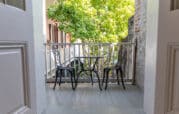 Two metal chairs and a small table on a porch overlooking a tree, with open doors framing the view.