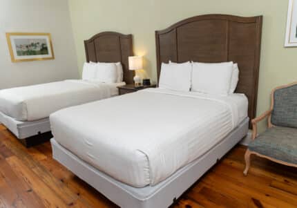 Hotel room with two neatly made double beds, each with white linens and wooden headboards. A nightstand with a lamp is between the beds. A framed picture hangs on the wall. A chair is to the right.