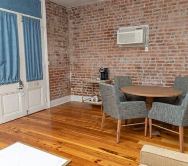 Room with brick walls, wooden floor, a round table with four blue chairs, air conditioner on the wall, and a small cart with a coffee machine by the window with blue curtains.