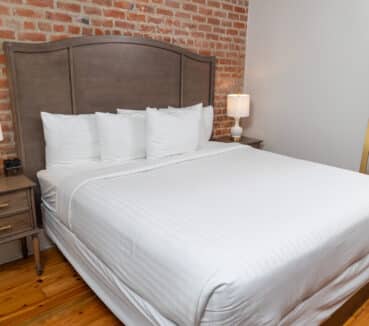 A neatly made bed with white linens, flanked by wooden nightstands and lamps. Exposed brick wall and a mirror on the right side.