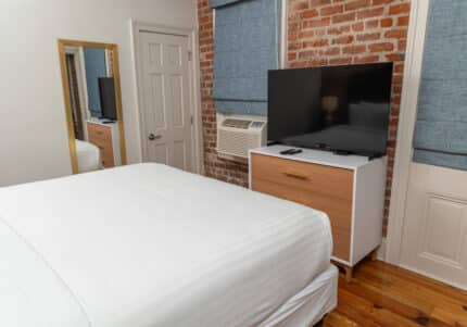 Bedroom with a bed, brick accent wall, mirror, dresser, TV, and window air conditioner. White bedding and wooden floor are visible.