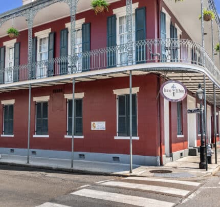 A red two-story building stands on a corner with ornate iron balconies and hanging plants. Signs read "Inn on St. Peter" and "St. Peter St." A crosswalk is visible in the foreground.