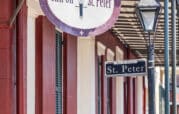 Street view of a red brick building with a sign reading "Inn on St. Peter" and a street sign for "St. Peter" in New Orleans. A vintage lamp post is visible next to the signs.