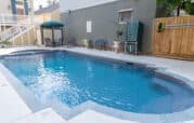 A small backyard swimming pool with clear water, surrounded by a concrete deck. There's patio furniture and a grill on the side, with a wooden fence and buildings in the background.