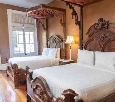 A room with two ornate wooden canopy beds, white bedding, a lamp between them, and large windows with light filtering through blinds.