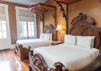 A room with two ornate wooden canopy beds, white bedding, a lamp between them, and large windows with light filtering through blinds.