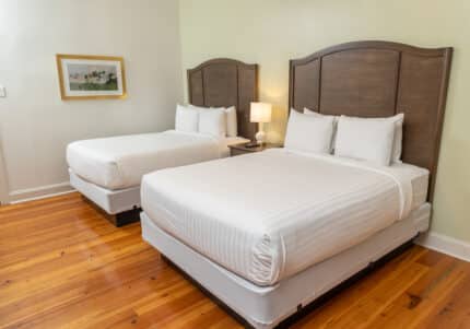 A hotel room with two double beds, white linens, a bedside lamp, and a framed picture on the wall. The floor is wooden.