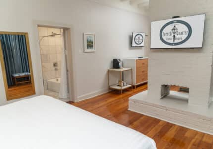 Hotel room with a bed, wall-mounted TV, dresser, mirrored bathroom door, and a coffee maker on a small table. Floors are wooden, and walls are light-colored.