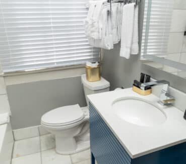 Small modern bathroom with a toilet, sink, and mirror. Towels hang on a rack above the toilet. Toiletries are arranged on the sink counter. A window with blinds is above the toilet.