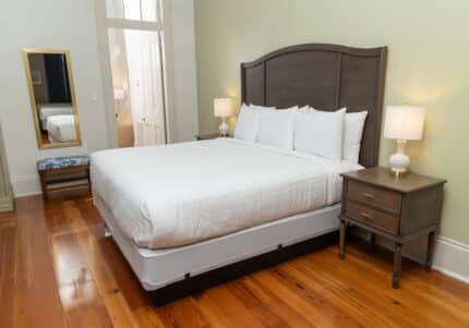 A neatly made king-sized bed with white linens and pillows, flanked by two bedside tables and lamps, on a wooden floor with a large mirror and blue cushioned bench nearby.