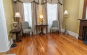 A room with wooden floors, two striped armchairs, a small wooden table, and three lamps in front of large windows with curtains. A decorative fireplace is on the right.