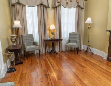 A room with wooden floors, two striped armchairs, a small wooden table, and three lamps in front of large windows with curtains. A decorative fireplace is on the right.