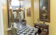 Yellow hallway with checkered floor, ornate chandeliers, and large paintings. Antique furniture includes a marble-top console and small seating area. Doorway visible at end.