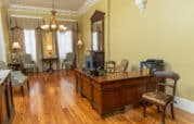 An elegant office with a wooden desk, a leather chair, and a printer. Two upholstered chairs and a small table sit by the window. Chandelier lights hang from the ceiling.
