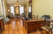 Office with a wooden desk, computer, chairs, and a printer. Elegant decor includes a chandelier, mirror, lamps, and striped chairs by the window. Light-colored walls and hardwood floor.