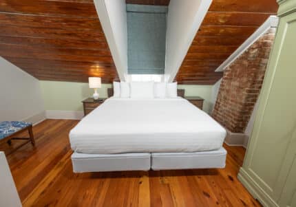 A cozy attic bedroom with a wooden ceiling, double bed, bedside lamp, and exposed brick wall. A window with a blue shade is centered above the bed.