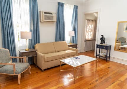 A living room with a beige sofa, blue armchair, wooden floor, coffee table, and two side tables with lamps. An air conditioner is above the sofa, and a mirror hangs on the wall.