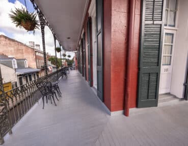 A long balcony with wrought iron railings, outdoor seating, and hanging plants. The building has red walls, green shutters, and a white door numbered 18.