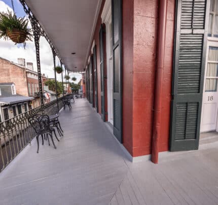 A long balcony with wrought iron railings, outdoor seating, and hanging plants. The building has red walls, green shutters, and a white door numbered 18.