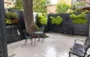 A small patio features a round table with two chairs, a bench, and a large tree. The black fence has hanging planters filled with greenery. The floor is light-colored and glossy.