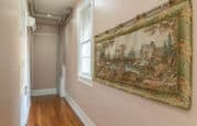 Narrow hallway with wooden floor, light beige walls, and a window. A decorative tapestry depicting a nature scene hangs on the right wall. Pipes and ductwork are visible near the ceiling.