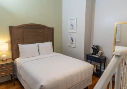 A neatly made bed with white linens is beside a wooden nightstand with a lamp. A small table holds a coffee machine. Two bird pictures hang on the pale green wall.