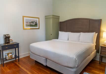 A neatly made double bed with white linens in a bedroom. A nightstand with a lamp is on one side, and a small table with a coffee maker is on the other side. A picture hangs on the wall.