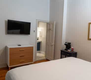 Hotel room with a wall-mounted TV, dresser, and a small table holding a coffee maker. Open door leads to a bathroom. A framed picture is on the wall above the table.