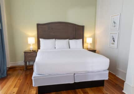 A neatly made bed with white linens and two pillows is flanked by two wooden nightstands. Each nightstand holds a lamp. Two framed pictures of birds hang on the adjacent wall.