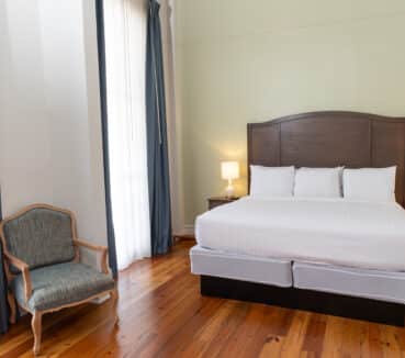 A hotel room with a large bed between two nightstands, each with a lamp. Blue curtains flank a window, and a wooden chair with a cushioned seat is in the corner.
