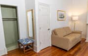 A room with a beige couch, floor lamp, wall art, and wooden stool with a blue cushion. An open closet and a full-length mirror are next to a closed white door. Wood flooring throughout.