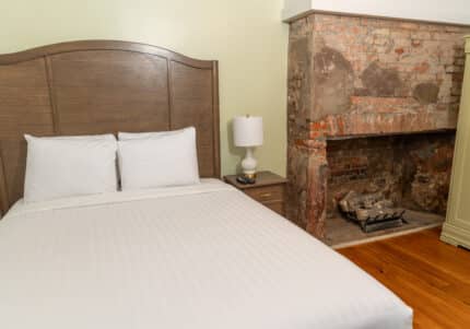 A bedroom with a double bed, wooden headboard, and white linens next to a nightstand holding a lamp. An old, brick fireplace is visible beside the bed.