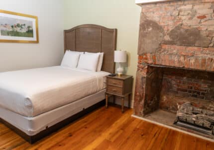 Bedroom with a neatly made bed, wooden headboard, a bedside table with a lamp, an exposed brick fireplace, wooden floor, and a framed picture on the wall.