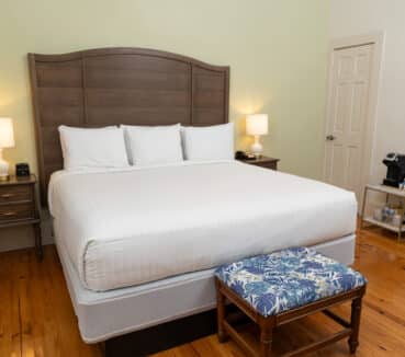Bedroom with a wooden floor, featuring a king-sized bed, two bedside tables with lamps, a small bench at the foot of the bed, and a coffee maker on a stand in the corner.