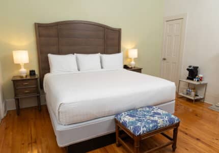 Bedroom with a wooden floor, featuring a king-sized bed, two bedside tables with lamps, a small bench at the foot of the bed, and a coffee maker on a stand in the corner.